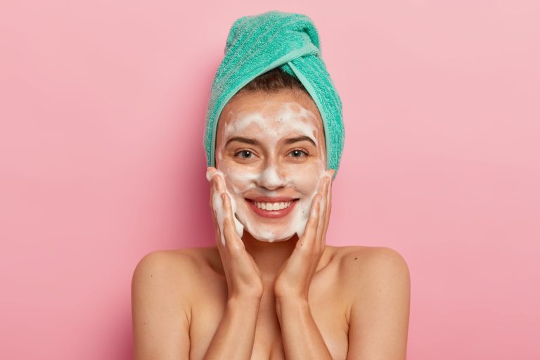 positive young female has toothy smile has perfect teeth pats skin with liquid sanitary soap washes with foaming gel wakes up morning having beauty routine